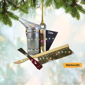 May include: A silver bee smoker with a chimney and a handle, decorated with colorful Christmas lights. The smoker is attached to a wooden brush with a brown handle and a green bristle brush. The brush is also decorated with Christmas lights. The ornament is personalized with the name "Emma".