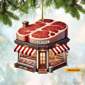 May include: A personalized Christmas ornament shaped like a butcher shop with a red and white striped awning. The shop is made of gingerbread and has a large, red, raw steak on top. The shop is labeled "JACKSON" and has a sign that says "PERSONALIZED".