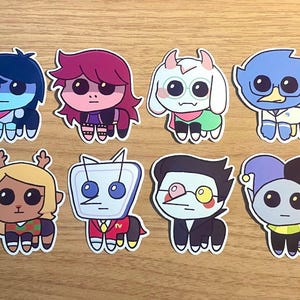 Puede incluir: Una colección de ocho pegatinas de personajes de dibujos animados. Cada pegatina presenta un diseño de personaje único con colores y características distintivas. Los personajes tienen ojos grandes y están de pie. Las pegatinas están sobre una superficie de madera.
