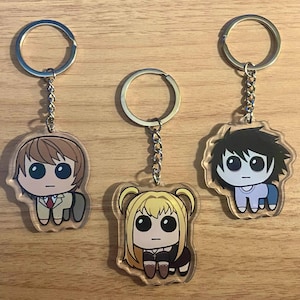 May include: Three anime-style character keychains. The keychains feature cartoon illustrations of characters with large eyes and unique outfits. Each keychain has a silver keyring and chain. The characters are on a wooden surface.