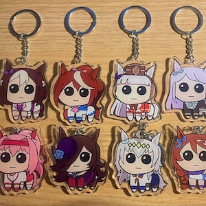 Może przedstawiać: Osiem akrylowych breloków do kluczy z postaciami z kreskówek przedstawiających konie z dużymi oczami i różnymi kolorami włosów i strojami. Każdy brelok ma srebrny łańcuszek i kółko. Postacie mają włosy w różnych kolorach, w tym brązowym, czerwonym, różowym i białym.