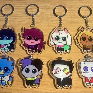 Könnte beinhalten: Acht Schlüsselanhänger aus Acryl mit Cartoon-Charakteren in verschiedenen Farben und Designs. Jeder Schlüsselanhänger hat eine silberne Kette und einen Ring. Zu den Charakteren gehören eine blau-schwarze Figur, eine Figur mit lila Haaren und eine weiß-grüne Figur.