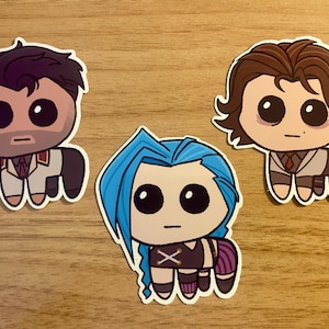 Op de afbeelding: Drie stickers met cartoonfiguren met grote ogen. De figuren zijn een man met bruin haar die een wit pak draagt, een vrouw met blauw haar die een zwart en paars outfit draagt, en een man met bruin haar die een wit pak draagt met een rode stropdas.