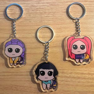 May include: Three acrylic keychains featuring cartoon characters with large eyes and various hairstyles. Each keychain has a silver-colored ring and chain. The characters are dressed in different outfits, with colors including purple, black, pink, yellow, and blue.