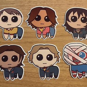 Peut inclure: Six autocollants de personnages de dessins animés avec des expressions et des vêtements différents. Les personnages portent tous des vêtements de couleurs différentes et ont des coiffures différentes. Les autocollants sont tous sur une surface en bois.