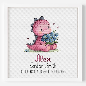 May include: A framed cross-stitch featuring a pink dinosaur holding blue flowers. The text "Alex Jordan Smith" is below the dinosaur, with the date, time, and measurements: 21 in., 1 lb. 10 oz.