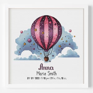 Op de afbeelding: Ingelijste kruissteekkunst met een roze en paarse luchtballon met sterren, boven een blauw-wit wolkenlandschap. De tekst luidt "Anna Marie Smith" met geboortedetails: 53 cm, 737 gram. De lijst is wit.