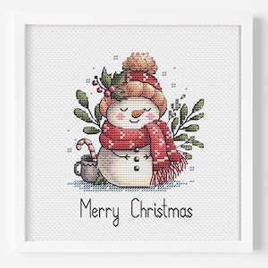 May include: A cross-stitch pattern featuring a snowman wearing a red scarf and hat, holding a candy cane and a cup of hot cocoa. The snowman is surrounded by green leaves and the text "Merry Christmas" is below the snowman.