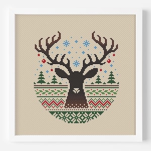 以下が含まれることがあります： 大きな角を持つトナカイ、雪の結晶、クリスマスツリーのモチーフが特徴の、額装されたクロスステッチのデザイン。デザインは、ベージュの背景に茶色、緑、赤、青の色合いを使用しています。アートワークは白いフレームで提示されています。