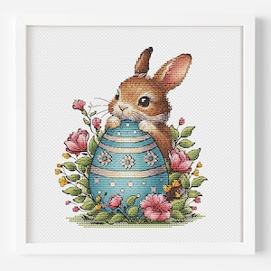 Puede incluir: Un conejo marrón sosteniendo un huevo de Pascua azul y blanco con un diseño floral. El conejo está rodeado de flores rosas y blancas.