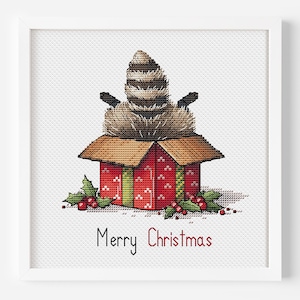 May include: Cross-stitch art of a raccoon emerging from a red gift box, decorated with green holly and berries. The box has a green ribbon and a repeating pattern. The words "Merry Christmas" are written in red below the box. The artwork is framed in a white square frame.