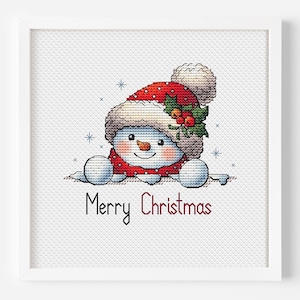 May include: A framed cross-stitch artwork featuring a smiling snowman wearing a red hat with white trim and holly. The snowman is wearing a red scarf and the words "Merry Christmas" are written below in red.