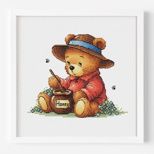 以下が含まれることがあります： 青いリボンと赤いシャツを着た、茶色の帽子をかぶったテディベアの額入りクロスステッチ。クマは「Honey」と書かれたポットの上でハニーディッパーを持っています。クマの周りをミツバチが飛んでいます。