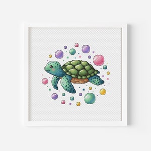 Może przedstawiać: Haft krzyżykowy przedstawiający żółwia morskiego z zielonym pancerzem i niebieskimi płetwami, otoczonego kolorowymi bąbelkami. Żółw się uśmiecha, a grafika jest oprawiona w białą kwadratową ramę. Tło jest białe.