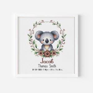 Op de afbeelding: Een witte ingelijste kruissteekborduursel met een schattige koalabeer die in een krans van groene bladeren en bruine bloemen zit. De tekst "Jacob Thomas Smith 01-01-2022 1:10pm | 21 in, 17.10 oz." staat onder de koala.