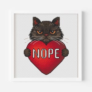 Op de afbeelding: Kruissteekkunst van een zwarte kat met oranje ogen en witte snorharen die een rood hart vasthoudt met het woord "NOPE" in wit. Het kunstwerk is ingelijst in een witte vierkante lijst.