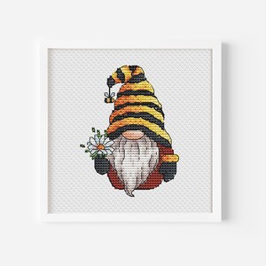 May include: A framed cross-stitch of a gnome holding a daisy bouquet. The gnome wears a striped yellow and black hat, a white beard, and a red and brown outfit. A small bee is on the hat. The artwork is displayed in a white frame.