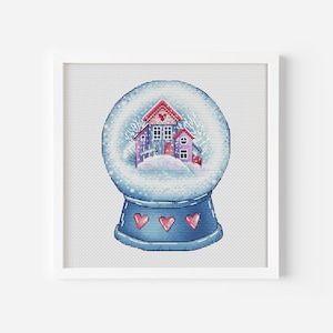 Könnte beinhalten: Eine blaue Schneekugel mit einem weißen Sockel und drei roten Herzen. Im Inneren der Kugel befindet sich ein kleines rosa und violettes Haus mit einem roten Dach und weißen Verzierungen.