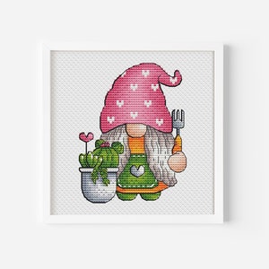 May include: A cross-stitch pattern of a gnome with a pink hat with white hearts, holding a gardening fork and a potted cactus plant.