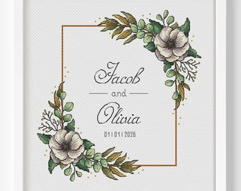 May include: A framed cross-stitch embroidery featuring a rectangular gold border decorated with floral arrangements of white flowers and green leaves. The center text reads Jacob and Olivia with the date 01 | 01 | 2026 in black script.
