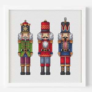 Puede incluir: Un patrón de bordado en punto de cruz con tres figuras de cascanueces en un marco blanco. La figura de la izquierda viste de verde, la del centro de rojo y la de la derecha de azul. Cada cascanueces tiene un sombrero, uniforme y barba.