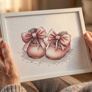 May include: A framed cross-stitch featuring two pink baby booties with large bows. The booties are detailed with shading and sit on a delicate, lacy design. The frame is white, and the artwork is held by a person.