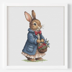 May include: A framed cross-stitch depicting a rabbit in a blue coat and red bow tie, carrying a basket of flowers. The rabbit stands on green grass. The artwork is presented in a white frame.