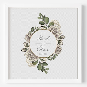 May include: A framed cross-stitch featuring a floral design with roses and greenery. The design includes a central circle with the names "Jacob and Olivia" and the date 01/01/2026. The frame is white.