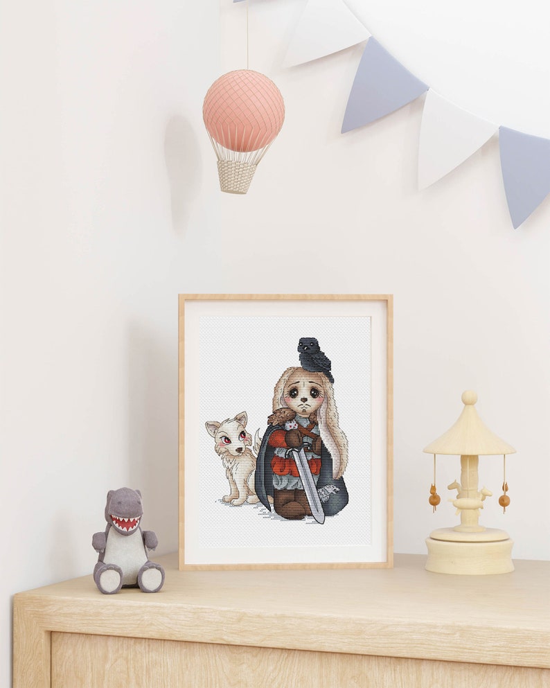 Puede incluir: Bordado de punto de cruz de un juguete de peluche vestido como un personaje de la serie de televisi&oacute;n Juego de Tronos. El juguete sostiene una espada y tiene un cuervo en la cabeza. Un perro de peluche blanco est&aacute; a su lado.