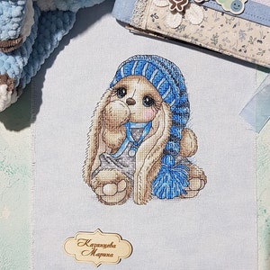 Puede incluir: Bordado de punto de cruz de un conejito marr&oacute;n que lleva un gorro y una bufanda a rayas azules y blancas. El conejito sostiene una pelota a rayas azules y blancas. El bordado est&aacute; firmado "Kazantseva Marina".