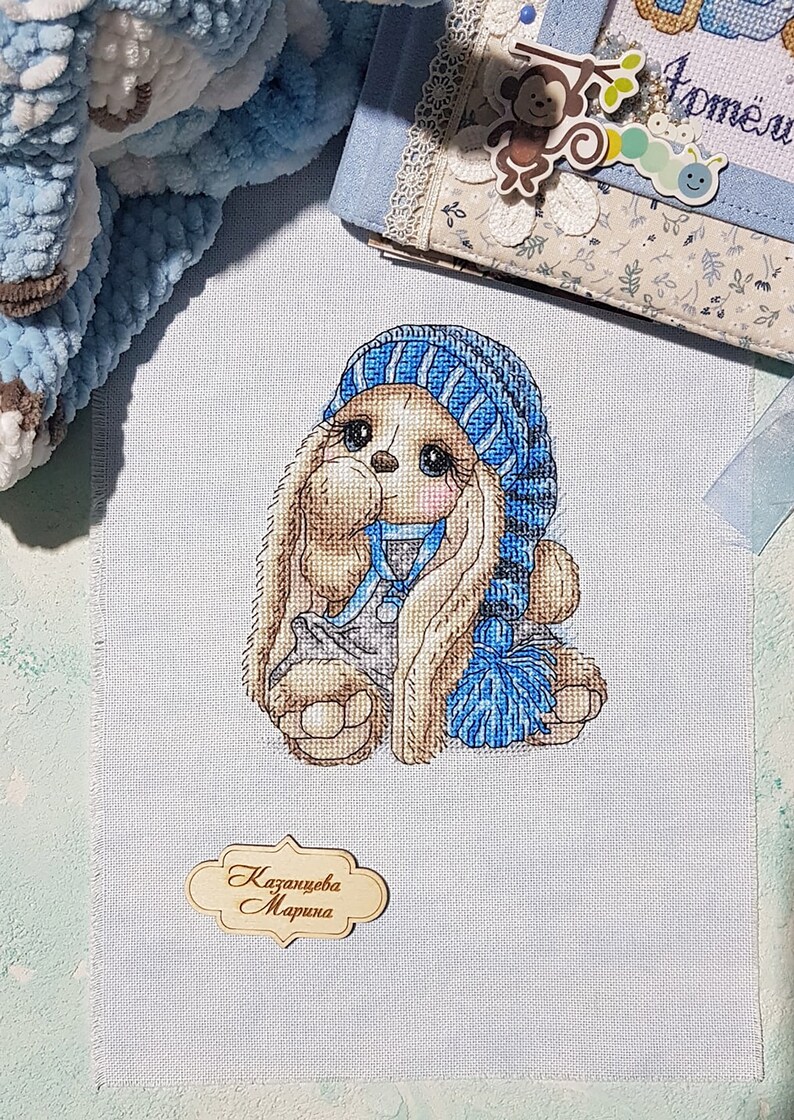 Puede incluir: Bordado de punto de cruz de un conejito marr&oacute;n que lleva un gorro y una bufanda a rayas azules y blancas. El conejito sostiene una pelota a rayas azules y blancas. El bordado est&aacute; firmado "Kazantseva Marina".