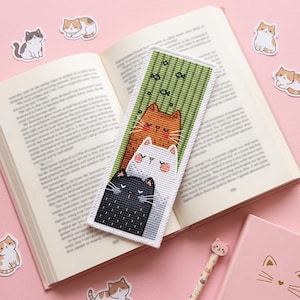 May include: A rectangular fabric bookmark featuring a cross-stitch design of three stacked cats in orange, white, and black. The background is a textured green with small black fish icons. The bookmark has a white stitched border and is placed on an open book.