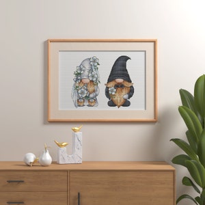 May include: A framed cross-stitch artwork featuring two gnomes, one dressed in white with flowers and the other in black with a beard. The gnomes are standing side-by-side and smiling.