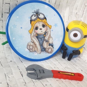 K&ouml;nnte beinhalten: Ein blauer Stickrahmen mit einem Kreuzstich-Design eines Hasen, der einen gelben Hut und eine Schutzbrille tr&auml;gt. Der Hase sitzt auf einem blauen Hintergrund mit wei&szlig;en Sternen. Ein gelbes Minion-Spielzeug und ein rot-grauer Schraubenschl&uuml;ssel sind ebenfalls auf dem Bild zu sehen.