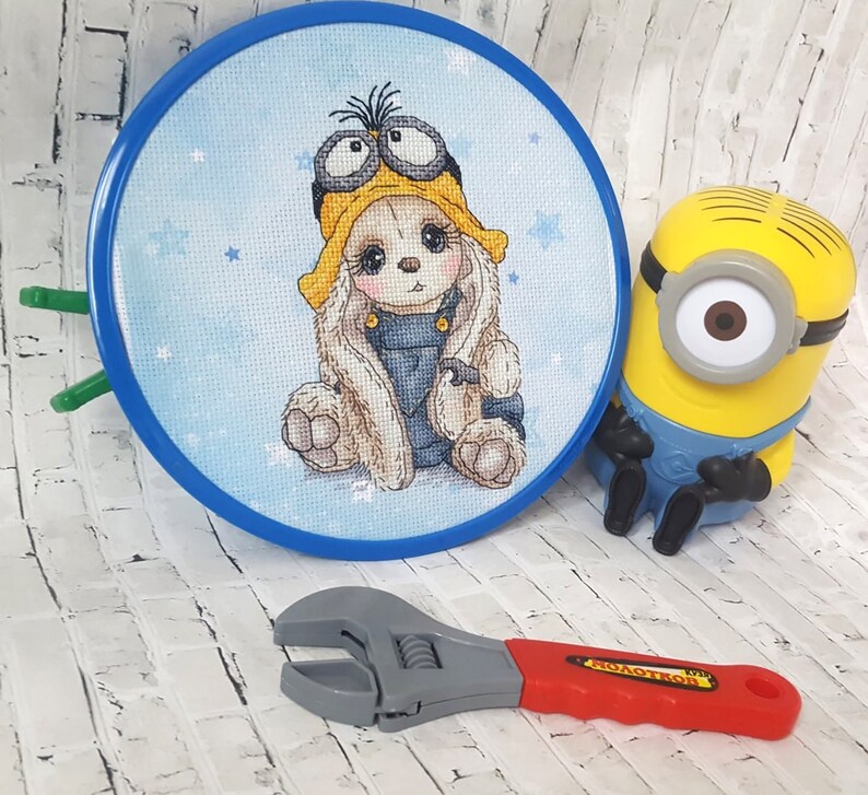 K&ouml;nnte beinhalten: Ein blauer Stickrahmen mit einem Kreuzstich-Design eines Hasen, der einen gelben Hut und eine Schutzbrille tr&auml;gt. Der Hase sitzt auf einem blauen Hintergrund mit wei&szlig;en Sternen. Ein gelbes Minion-Spielzeug und ein rot-grauer Schraubenschl&uuml;ssel sind ebenfalls auf dem Bild zu sehen.