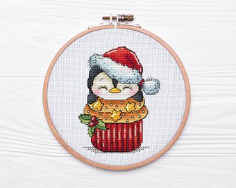 May include: A cross-stitch embroidery hoop with a penguin wearing a red Santa hat and a yellow and orange sweater with stars. The penguin is inside a red and white cupcake with green holly leaves.