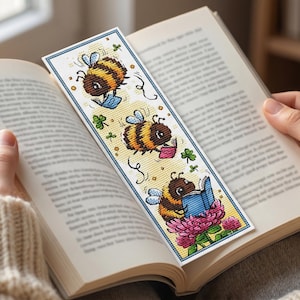 Op de afbeelding: Een kleurrijke boekenlegger met drie stripfiguren van bijen die boeken lezen. De bijen hebben bruin en geel gestreepte lichamen en zijn omringd door groene klavers. De boekenlegger ligt in een open boek, vastgehouden door iemand met een crèmekleurige trui.