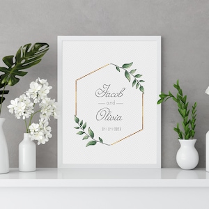 May include: A white framed print with a gold geometric frame and green foliage. The text on the print reads "Jacob and Olivia 01.01.2023".