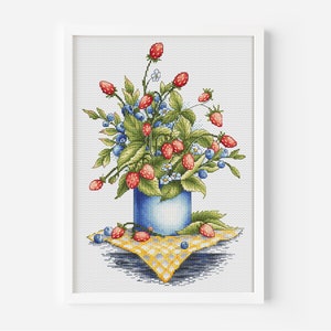 May include: A cross-stitch embroidery of a blue vase filled with red and green strawberries and blue blueberries. The vase is sitting on a yellow and white checkered tablecloth.