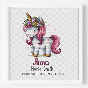 May include: A framed cross-stitch depicting a unicorn with a pink mane and tail, wearing a floral crown. The name "Anna Marie Smith" is stitched below, along with birth details: 53 cm, 737 g.
