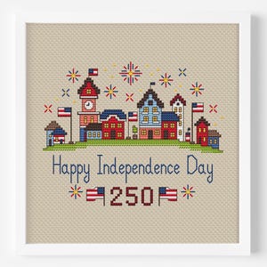 May include: Cross-stitch artwork featuring a patriotic town scene with buildings, flags, and fireworks. The text "Happy Independence Day" is displayed, along with "250" and decorative elements. The artwork is framed in a white border.