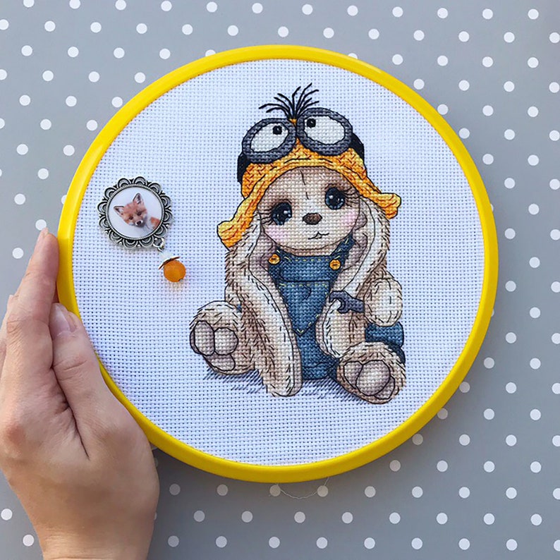 K&ouml;nnte beinhalten: Eine Kreuzstich-Stickerei eines niedlichen Hasen, der einen gelben Hut mit Schutzbrille und einen blauen Jeans-Overall tr&auml;gt. Der Hase sitzt auf einem wei&szlig;en Hintergrund.