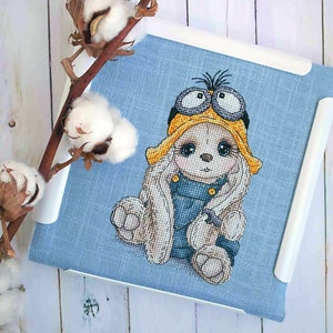 K&ouml;nnte beinhalten: Kreuzstich-Stickerei eines wei&szlig;en Kaninchens, das ein gelbes und schwarzes Minion-Kost&uuml;m mit Schutzbrille tr&auml;gt. Das Kaninchen sitzt auf einem blauen Stoffhintergrund.