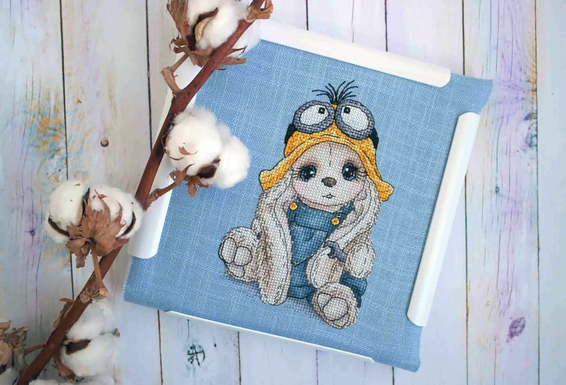 K&ouml;nnte beinhalten: Kreuzstich-Stickerei eines wei&szlig;en Kaninchens, das ein gelbes und schwarzes Minion-Kost&uuml;m mit Schutzbrille tr&auml;gt. Das Kaninchen sitzt auf einem blauen Stoffhintergrund.