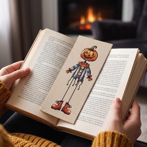 Peut inclure: Un marque-page rectangulaire en point de croix représentant un épouvantail à tête de citrouille sculptée, portant une chemise rayée bleue et blanche avec des boutons orange, un col à volants et des bottes orange sur un tissu beige clair.