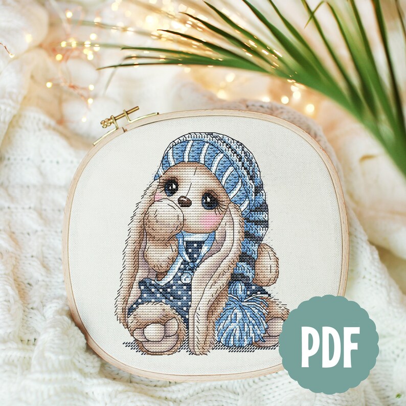 Puede incluir: Patr&oacute;n de punto de cruz de un conejito marr&oacute;n que lleva un gorro a rayas azul y blanco y un jersey de lunares azul y blanco. El conejito sostiene un pomp&oacute;n azul.