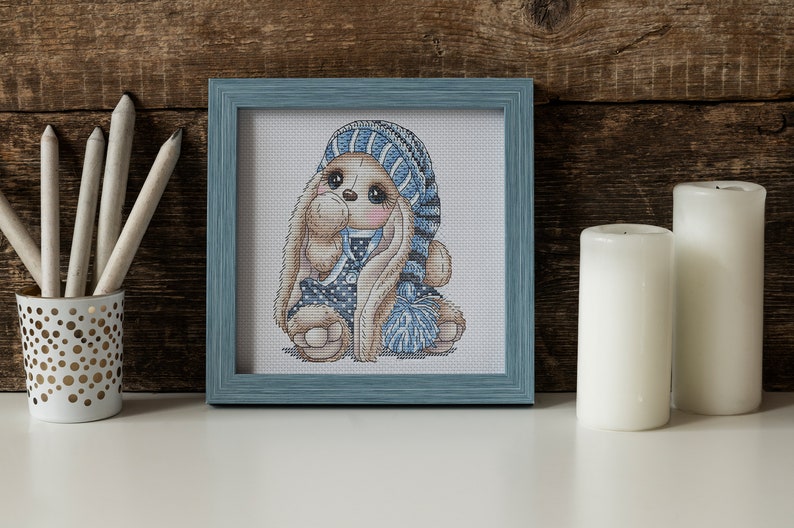 Puede incluir: Una bordado de punto de cruz de un conejo marr&oacute;n que lleva un gorro de punto azul y blanco y un vestido azul con lunares blancos. El conejo sostiene una bola de lana azul y blanca.