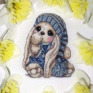 Puede incluir: Un patr&oacute;n de punto de cruz de un lindo conejito con un gorro de punto azul y blanco y un vestido de lunares azules y blancos. El conejito sostiene un pomp&oacute;n azul y blanco. El conejito est&aacute; rodeado de narcisos amarillos.