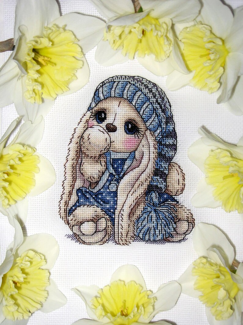 Puede incluir: Un patr&oacute;n de punto de cruz de un lindo conejito con un gorro de punto azul y blanco y un vestido de lunares azules y blancos. El conejito sostiene un pomp&oacute;n azul y blanco. El conejito est&aacute; rodeado de narcisos amarillos.