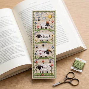 Può includere: Segnalibro fatto a mano a punto croce con quattro disegni di pecore, la parola «Baa», fiori, un sole e nuvole. Il segnalibro ha un bordo verde. Una bobina di filo verde e piccole forbici sono accanto al segnalibro, accanto a un libro aperto.